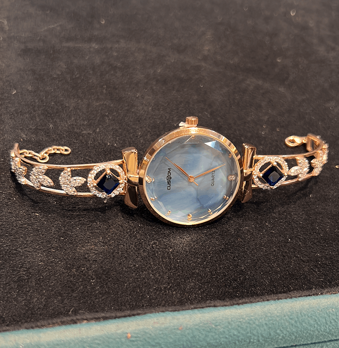 Rose gold women wristwatch with a beautiful band rose gold with a blue dial and 3 side leaf design on the band with 2 beautiful blue stones on either side of the band.