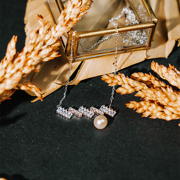 Elfy 925 Sterling Silver geometric necklace for women featuring a modern bar of baguette cut crystals and a single hanging round pearl. Artistically styled on a dark background with gold accents and wheat stalks, A sophisticated and unique fine jewelry gift 