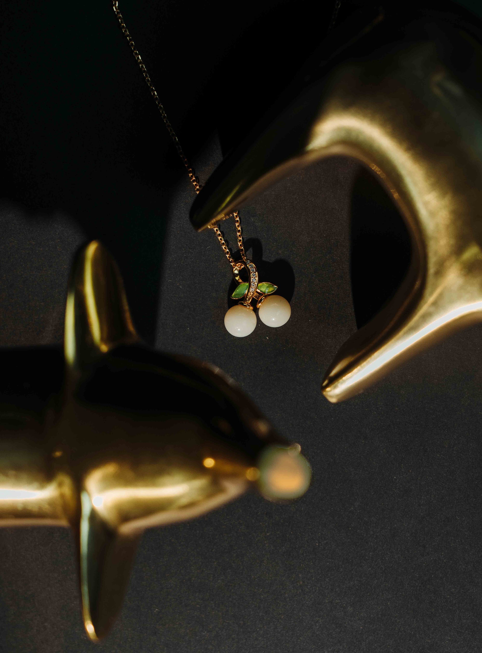 Gold-plated necklace featuring a cherry pendant made of two white round stones and green crystal leaves, artistically framed by golden dolphin figurines on a dark background—An elegant and whimsical jewelry gift for her.