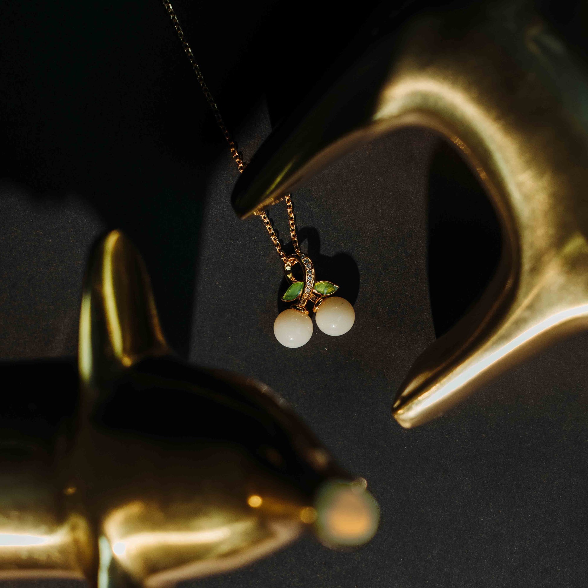 Gold-plated necklace featuring a cherry pendant made of two white round stones and green crystal leaves, artistically framed by golden dolphin figurines on a dark background—An elegant and whimsical jewelry gift for her.