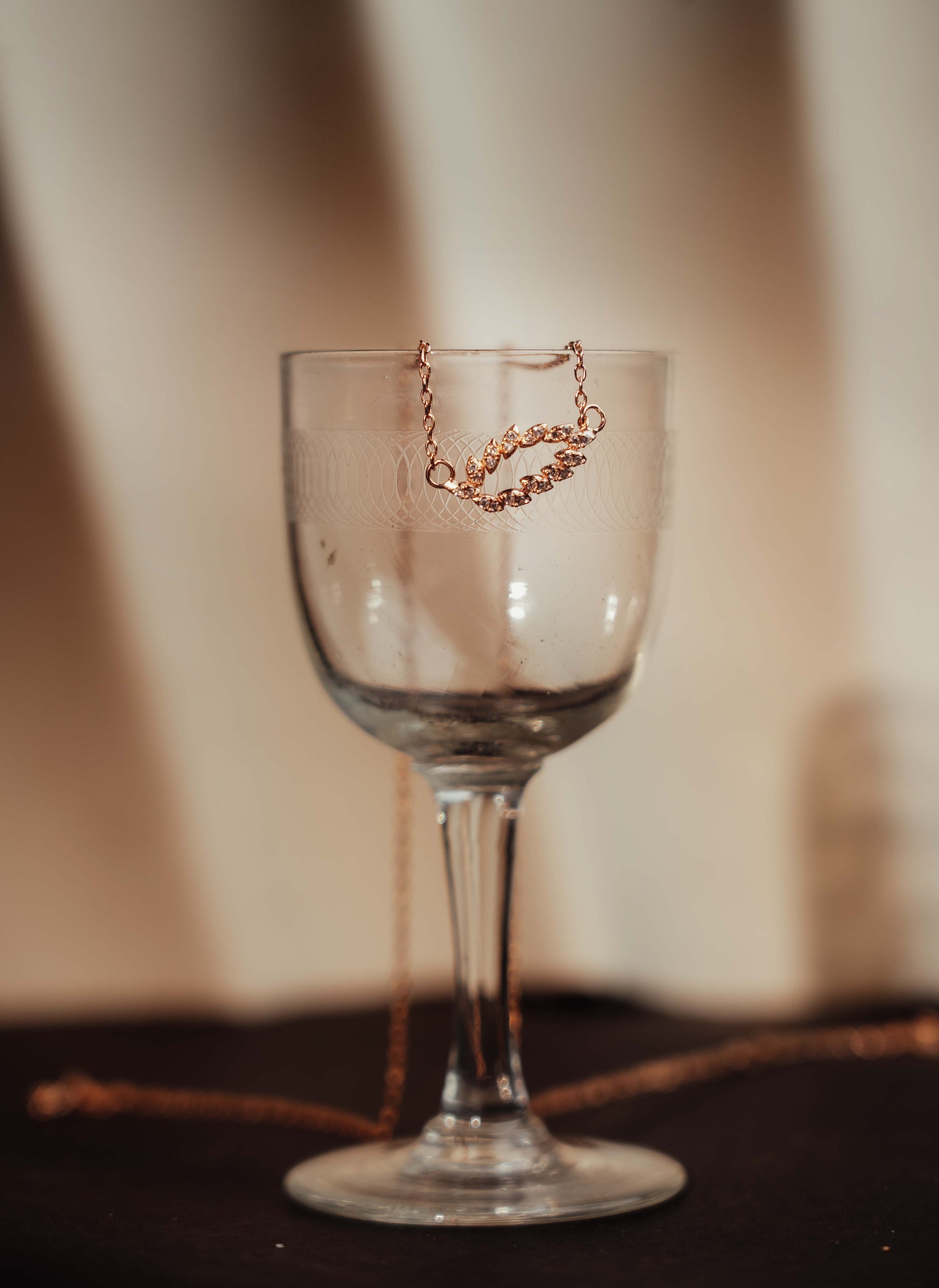 Elfy 925 Sterling Silver Rose Gold Leaf Pendant Necklace with shimmering marquise-cut crystals, artistically draped over a clear glass goblet in a warm studio setting—A premium and nature-inspired jewelry gift