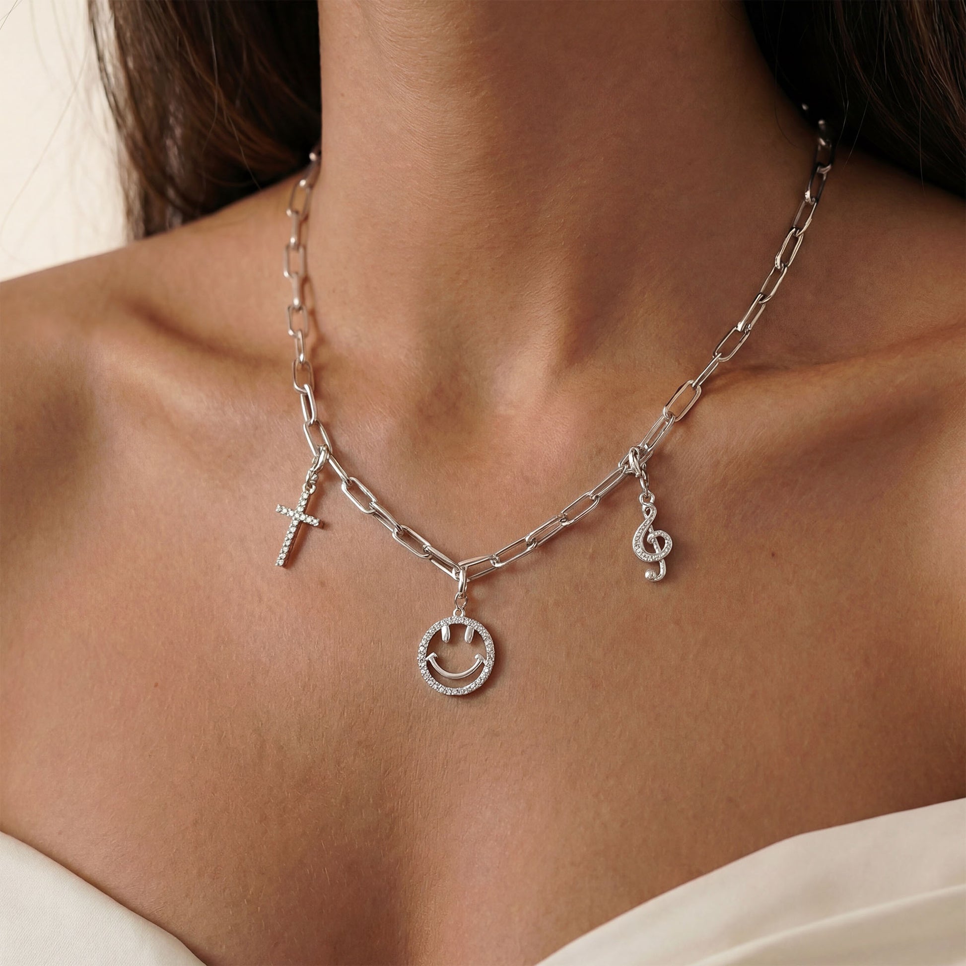 Paperclip charm chain with charms worn by a person, close-up view. The charms which are worn are smiley, sacred cross and music note charm all in silver